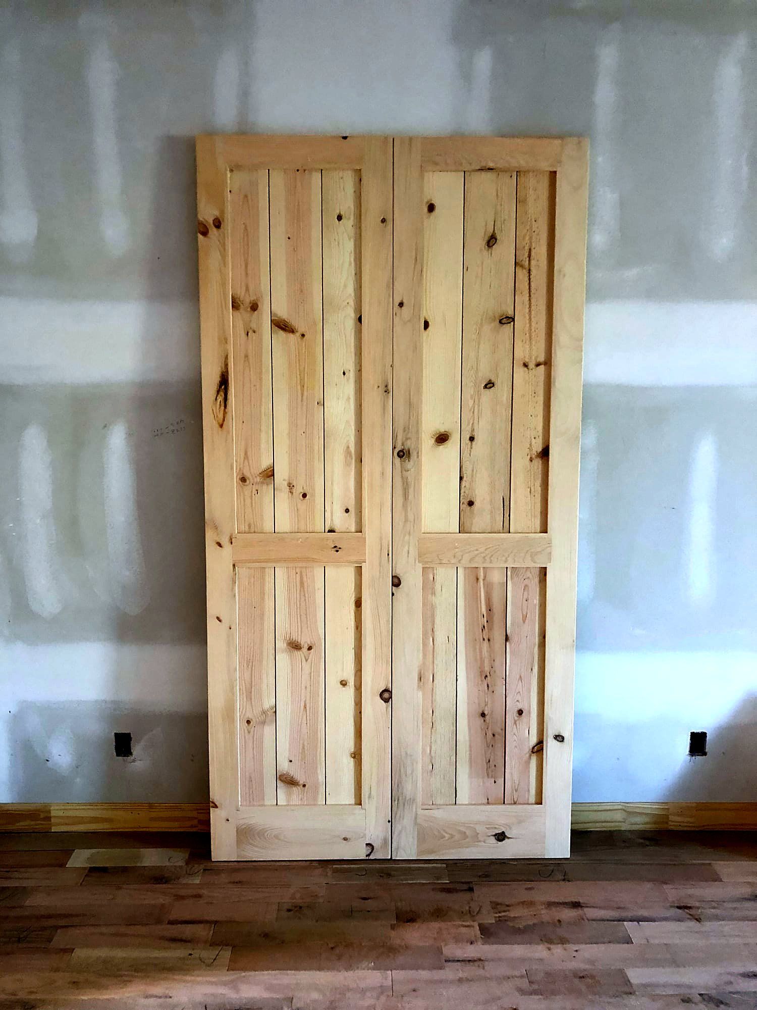 Pair of narrow two-panel knotty pine doors standing against drywall, natural finish with tight vertical grain and prominent knots