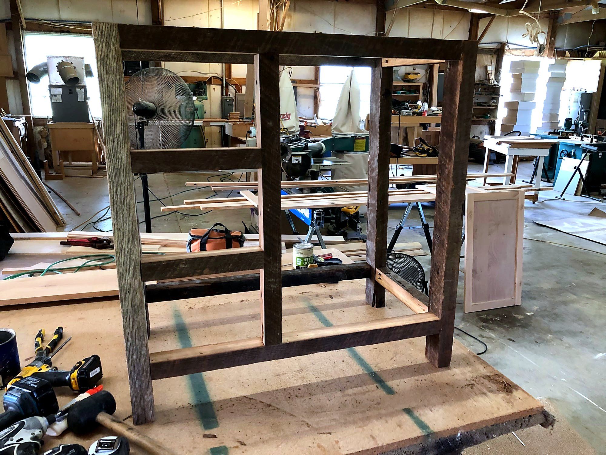 Reclaimed barn-wood frame being assembled on the shop floor, weathered gray uprights joined with mortise-and-tenon joints