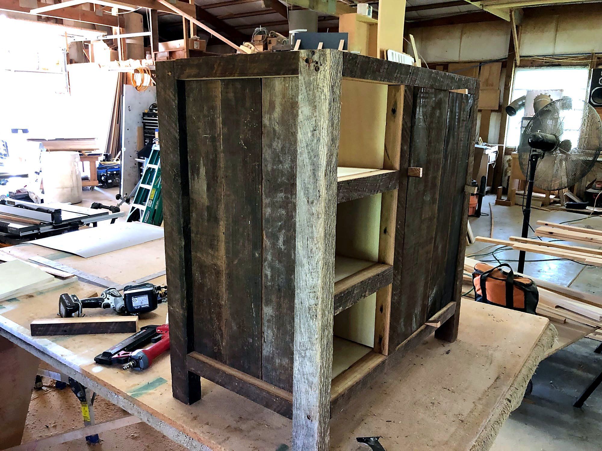 Cabinet carcass faced in weathered gray barn wood, mid-assembly on a shop workbench with open shelf bays and a hinged door