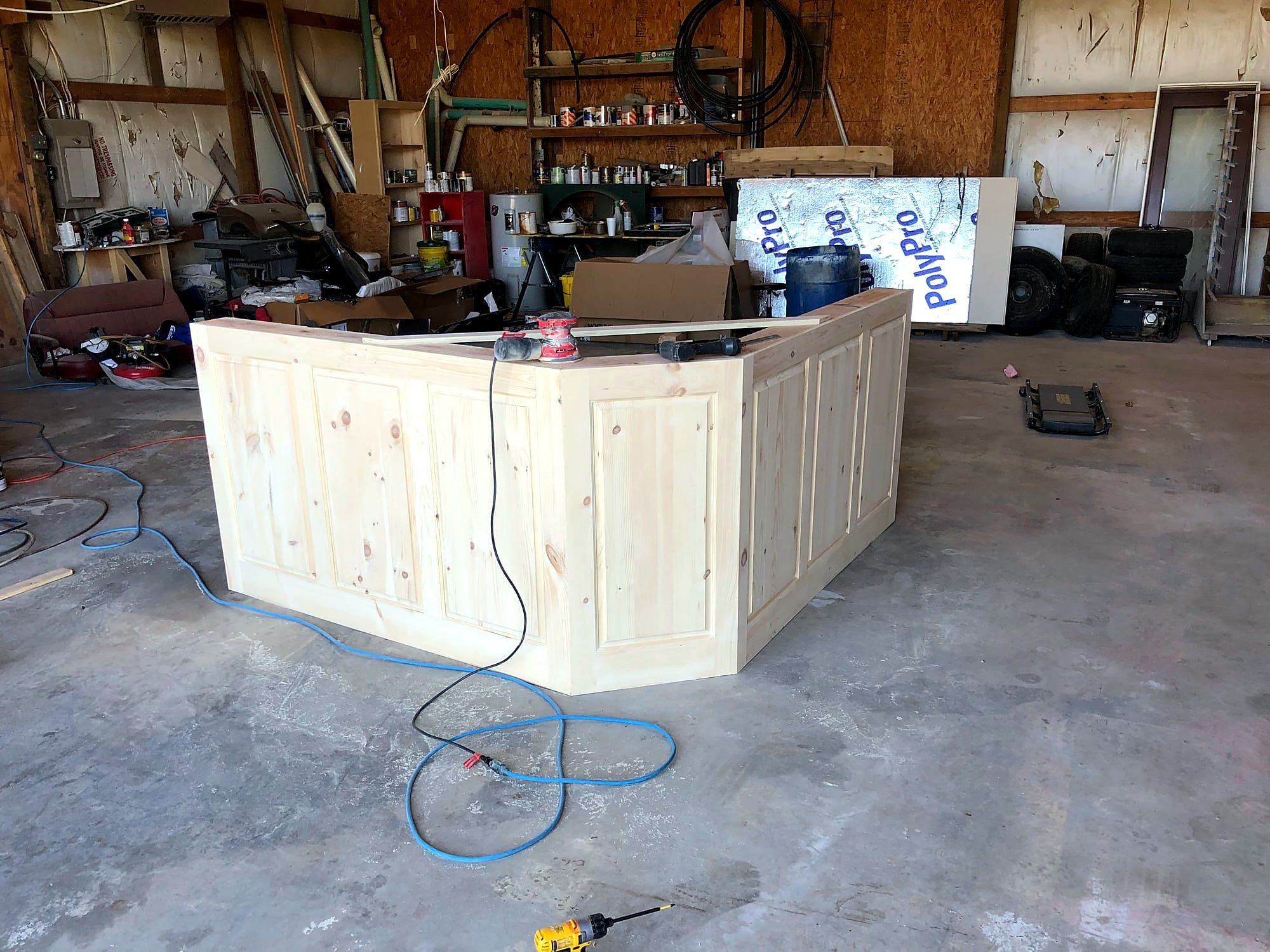 Knotty pine bar counter under construction, mitered raised-panel apron wrapping an angled corner, sander resting on the unfinished top
