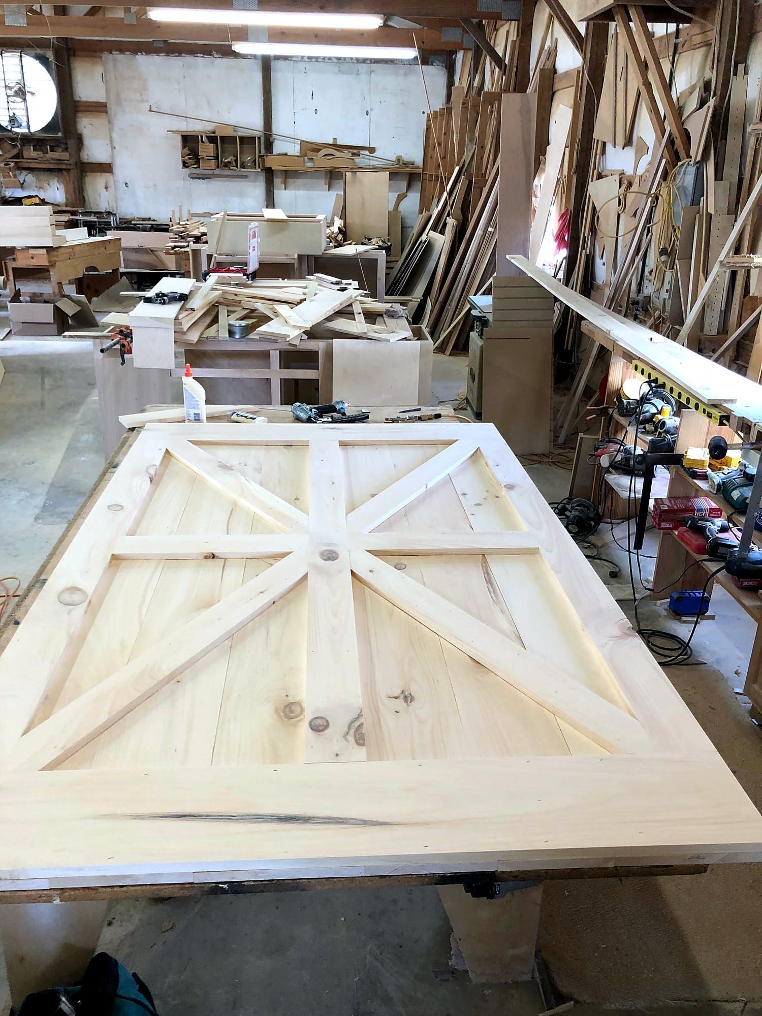 Large sliding door with a pine-plank face and a bold X-brace pattern spanning both halves, clamped and glued on a shop workbench