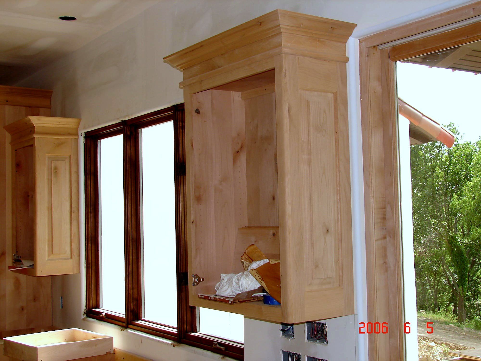 Knotty alder upper wall cabinet with a crown-topped face frame, door open, mounted beside a window near a covered porch entry