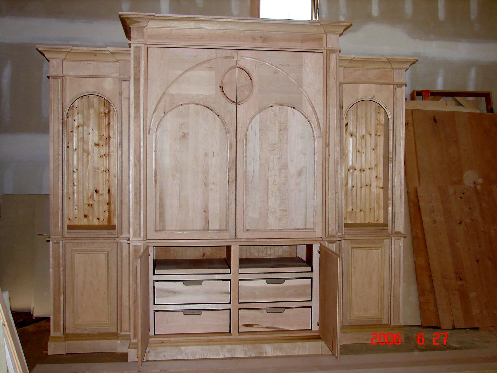 Ornate built-in wall unit with arched raised-panel doors, a circular medallion inset, and flanking fluted bookcase towers, unstained maple with open lower drawers