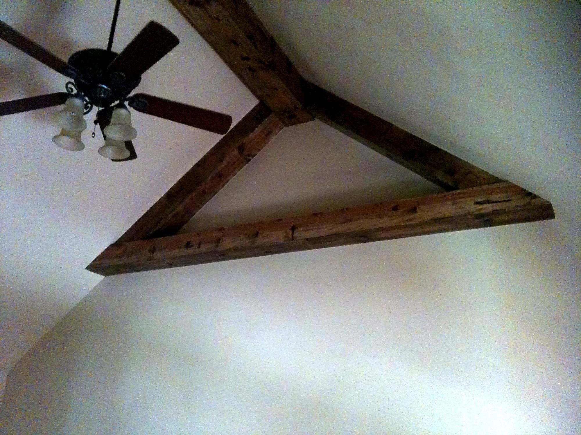 Ceiling fan mounted between exposed wood king-post truss beams on a plaster ceiling
